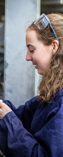 A female engineering student in navy-blue overalls selecting small metal components from a storage drawer, wearing safety goggles on her head. A female engineering student in navy-blue overalls selecting small metal components from a storage drawer, wearing safety goggles on her head.